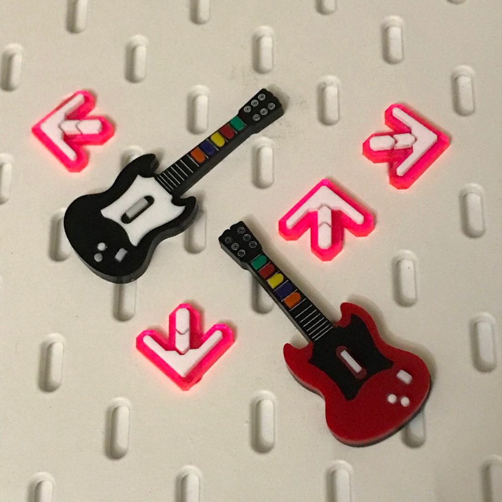 Photograph of plastic brooch pins. There is a black and red guitar, resembling the controllers from Guitar Hero complete with buttons along the fret bar. They're surrounded by four arrows from Dance Dance Revolution.